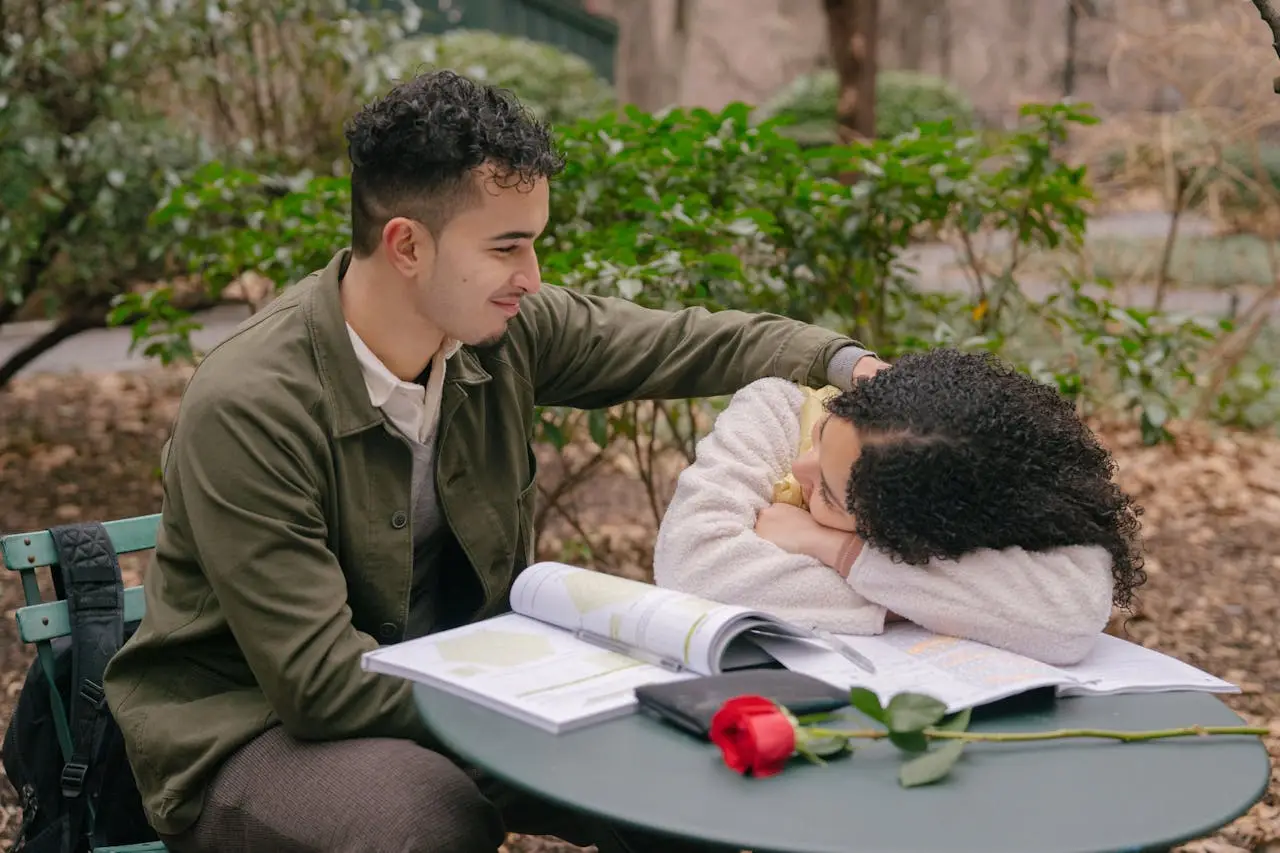 Cheerful ethnic couple smiling and looking at each other at table with textbook and bright red blooming rose
